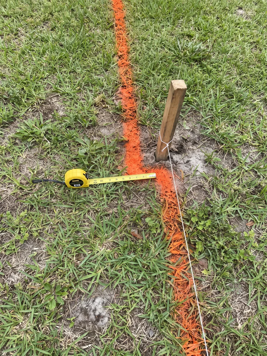 An orange marking-paint line turning a corner on a lawn, a wooden stake with a string line and a tape measure beside it