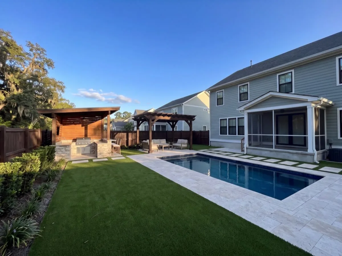 After: The same garden laid with artificial turf and travertine, with a pool deck, an outdoor kitchen and a pergola