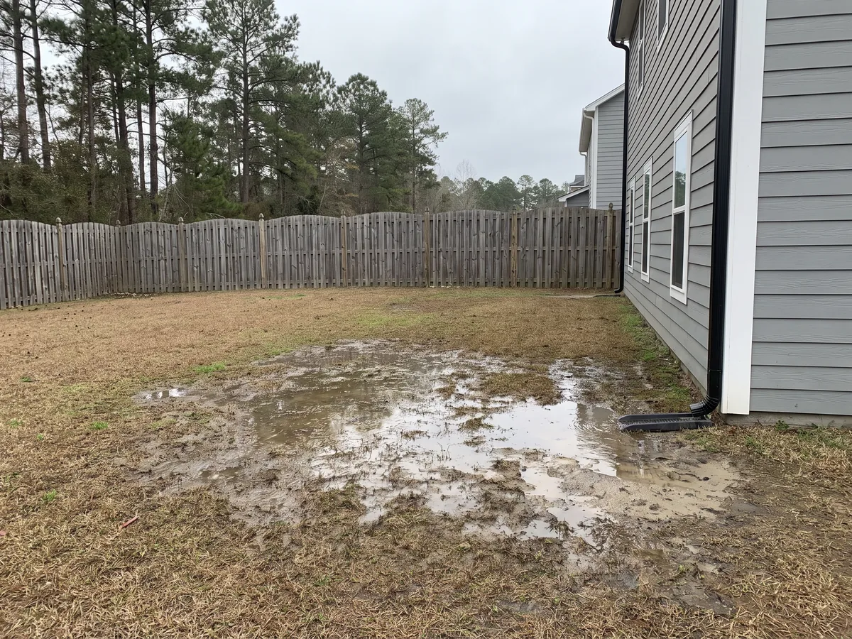 Before: A gray house's back yard flooded with standing muddy water in front of an old weathered fence