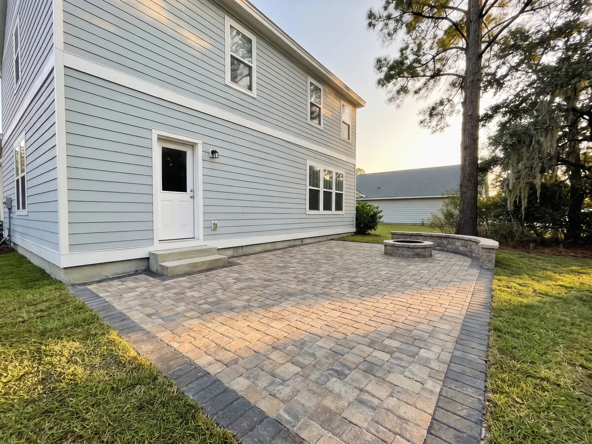 After: The same back yard with a tumbled paver patio in a charcoal border, a curved seating wall and a round stone fire pit